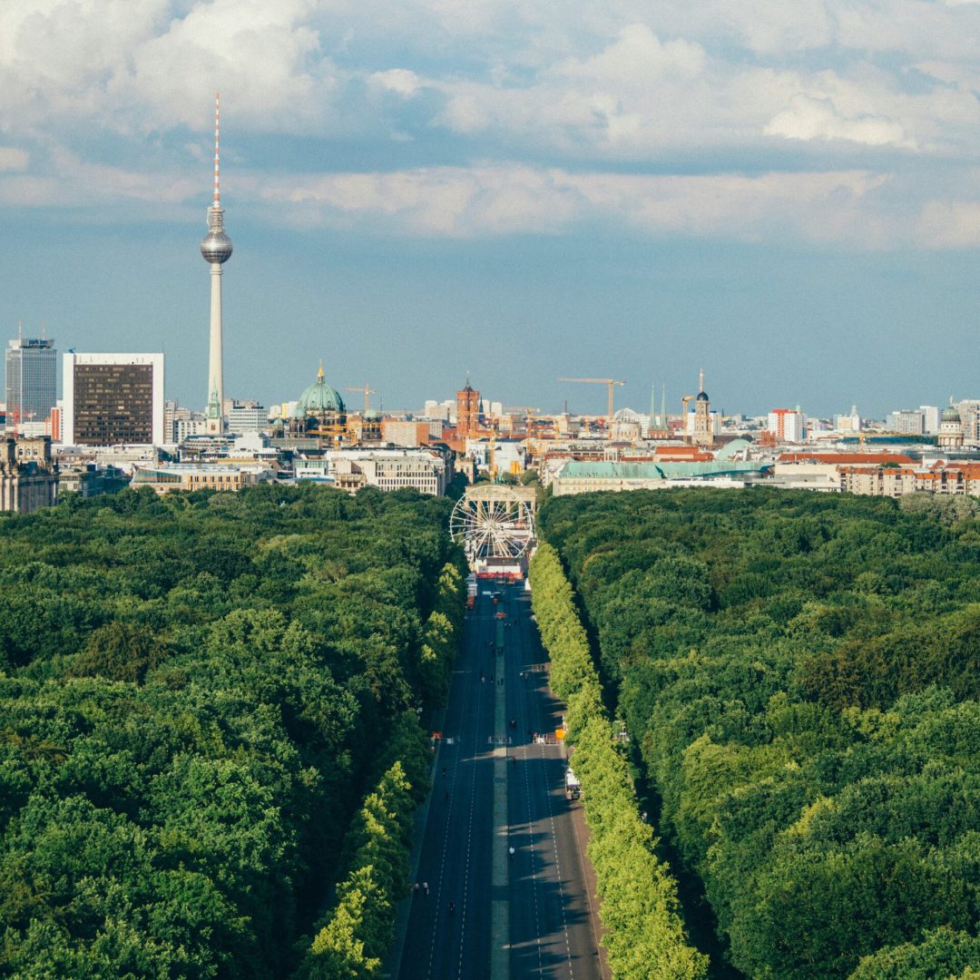 Blick über den Berliner Tiergarten mit der Straße des 17. Juni, die zentral durch den Park verläuft. Im Hintergrund sind der Berliner Fernsehturm, der Berliner Dom, das Rote Rathaus und weitere markante Gebäude der Hauptstadt zu sehen. Ein Riesenrad steht am Ende der Straße und befindet sich visuell vor der Berliner Skyline.