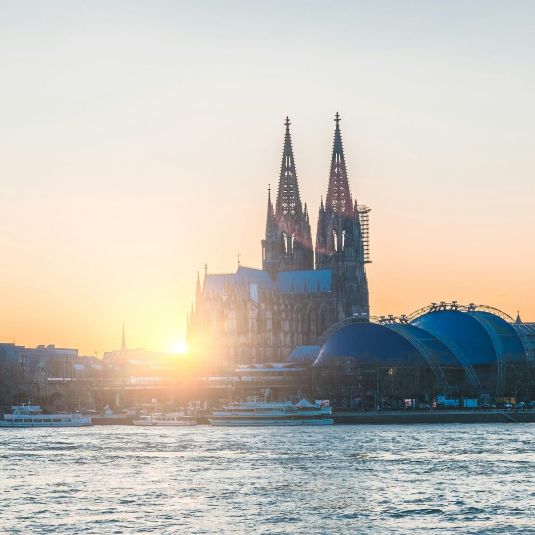 Das Bild zeigt den Kölner Dom vom Rhein aus. Der Rhein wird vor dem Kölner Dom präsentiert, dahinter zeigt sich der Musical Dome und der Bahnhof mit einigen Zügen. Im Hintergrund erstrahlt der Kölner Dom im Sonnenuntergang.