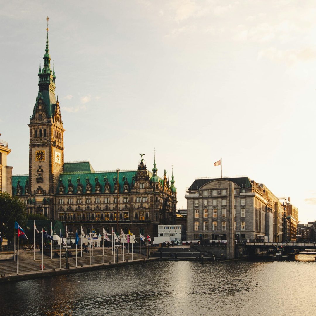 Das Bild zeigt das Hamburger Rathaus in der Altstadt von Hamburg. Davor befinden sich zahlreiche Fahnen an Flaggenmasten. Rechts verläuft die Alster.