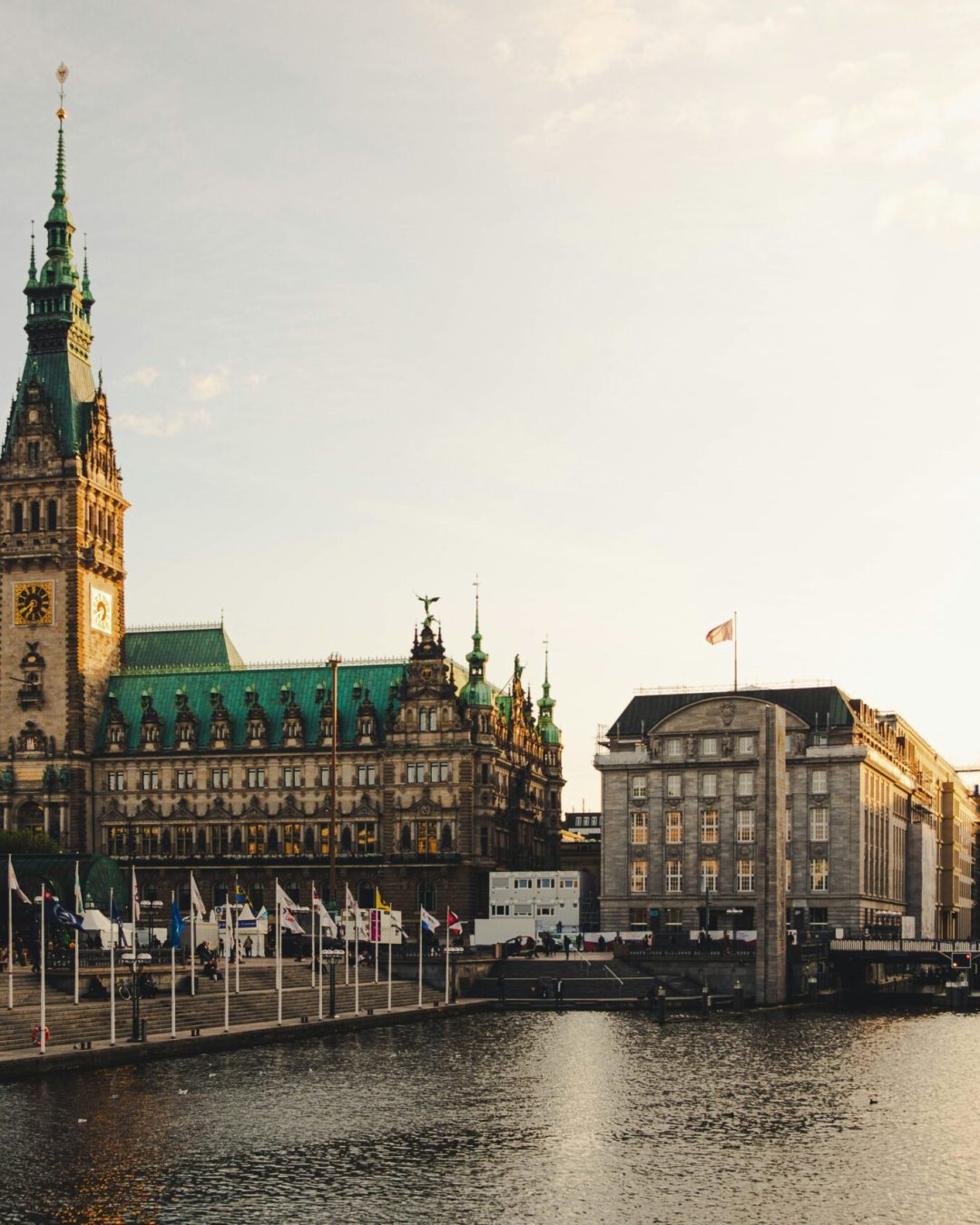 Das Bild zeigt das Hamburger Rathaus in der Altstadt von Hamburg. Davor befinden sich zahlreiche Fahnen an Flaggenmasten. Rechts verl&auml;uft die Alster.
